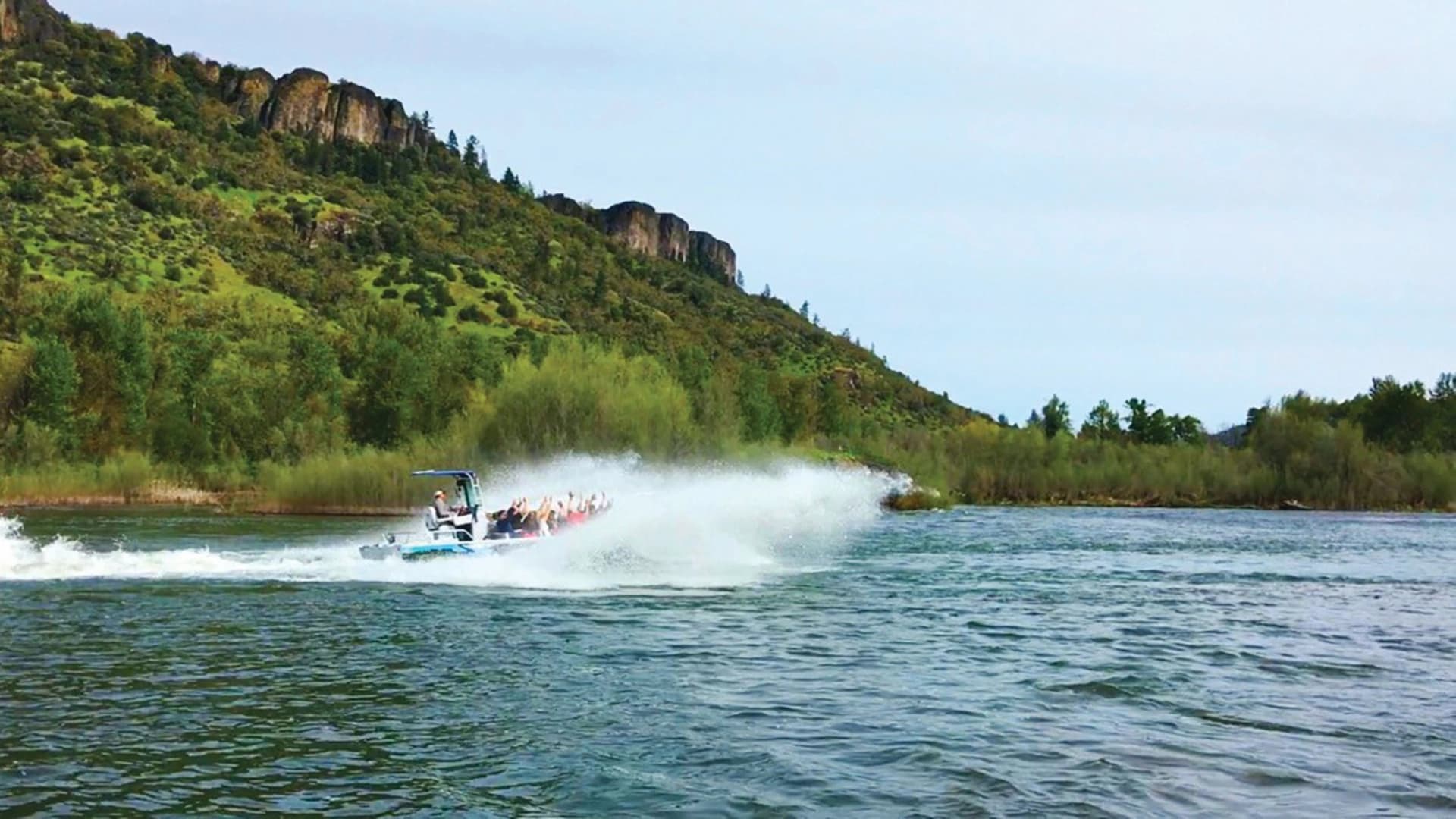 A speedboat creates a spray as it navigates a river surrounded by lush green hills.