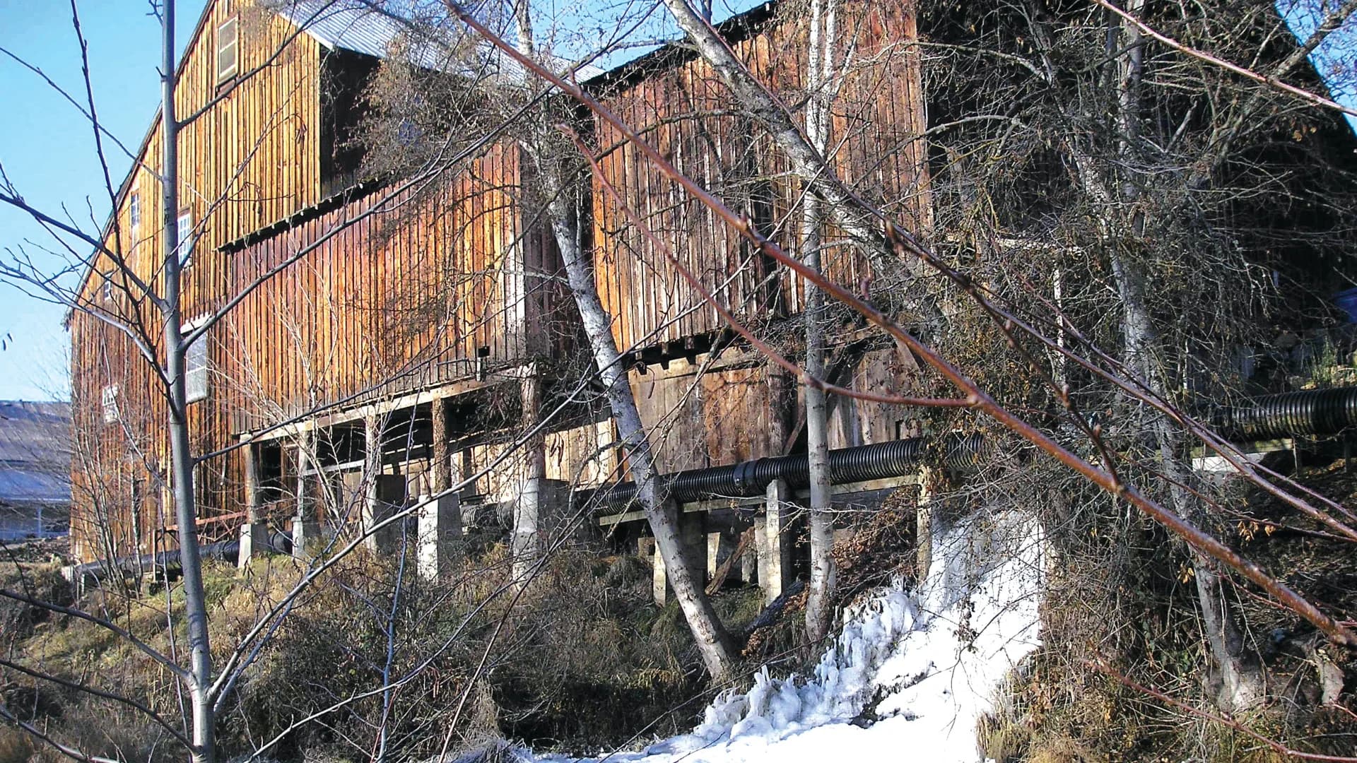 An old wooden building partially obscured by trees, with snow and a pipeline visible in the foreground.