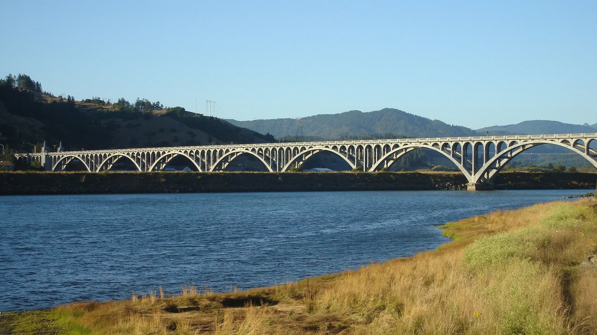 A large concrete arch bridge spans a river, with hills in the background and grassy banks in the foreground.