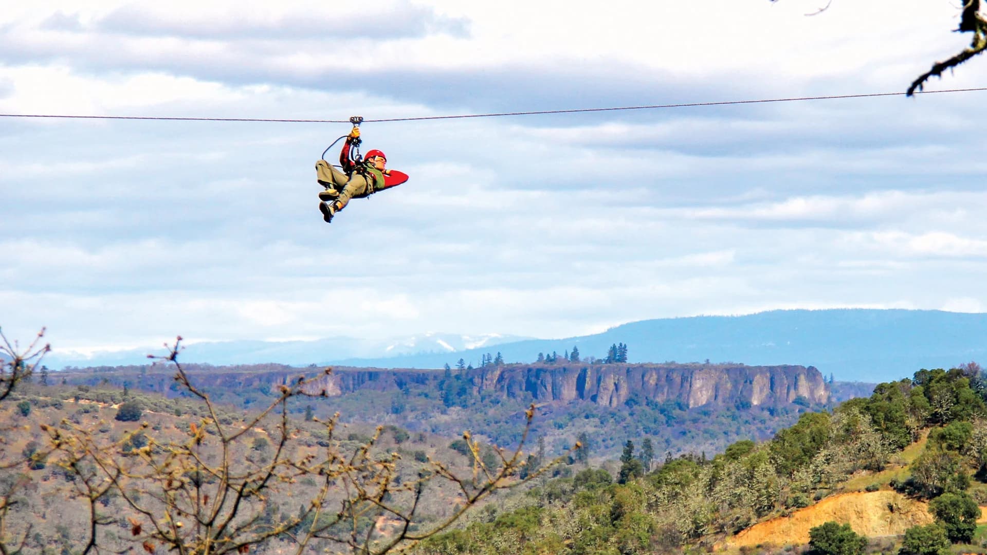 A person zip-lining over a mountainous landscape with cloudy skies in the background.