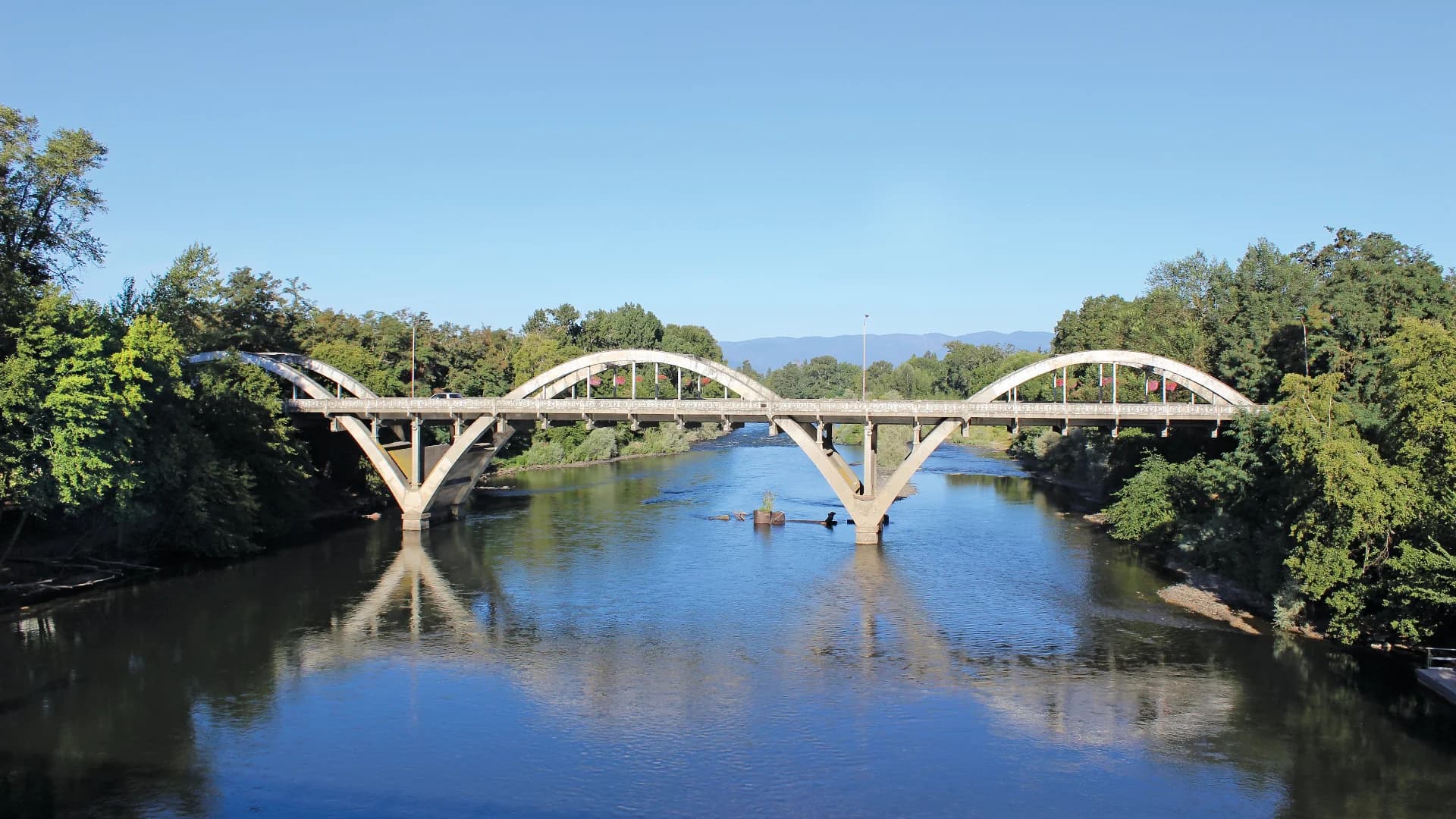 A large, arched concrete bridge spans a calm river, surrounded by greenery under a clear blue sky.