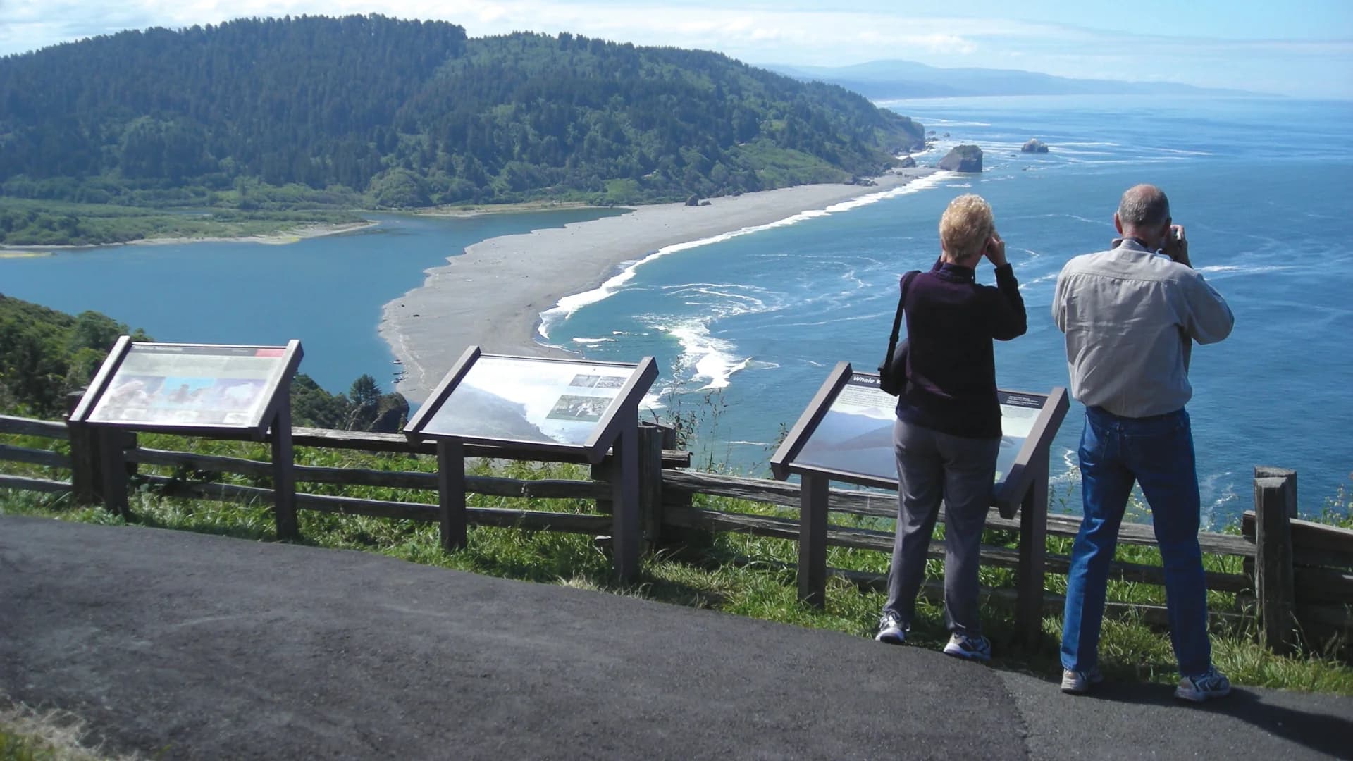 Two people stand on a viewpoint overlooking a beach and ocean, with information signs in front of them.