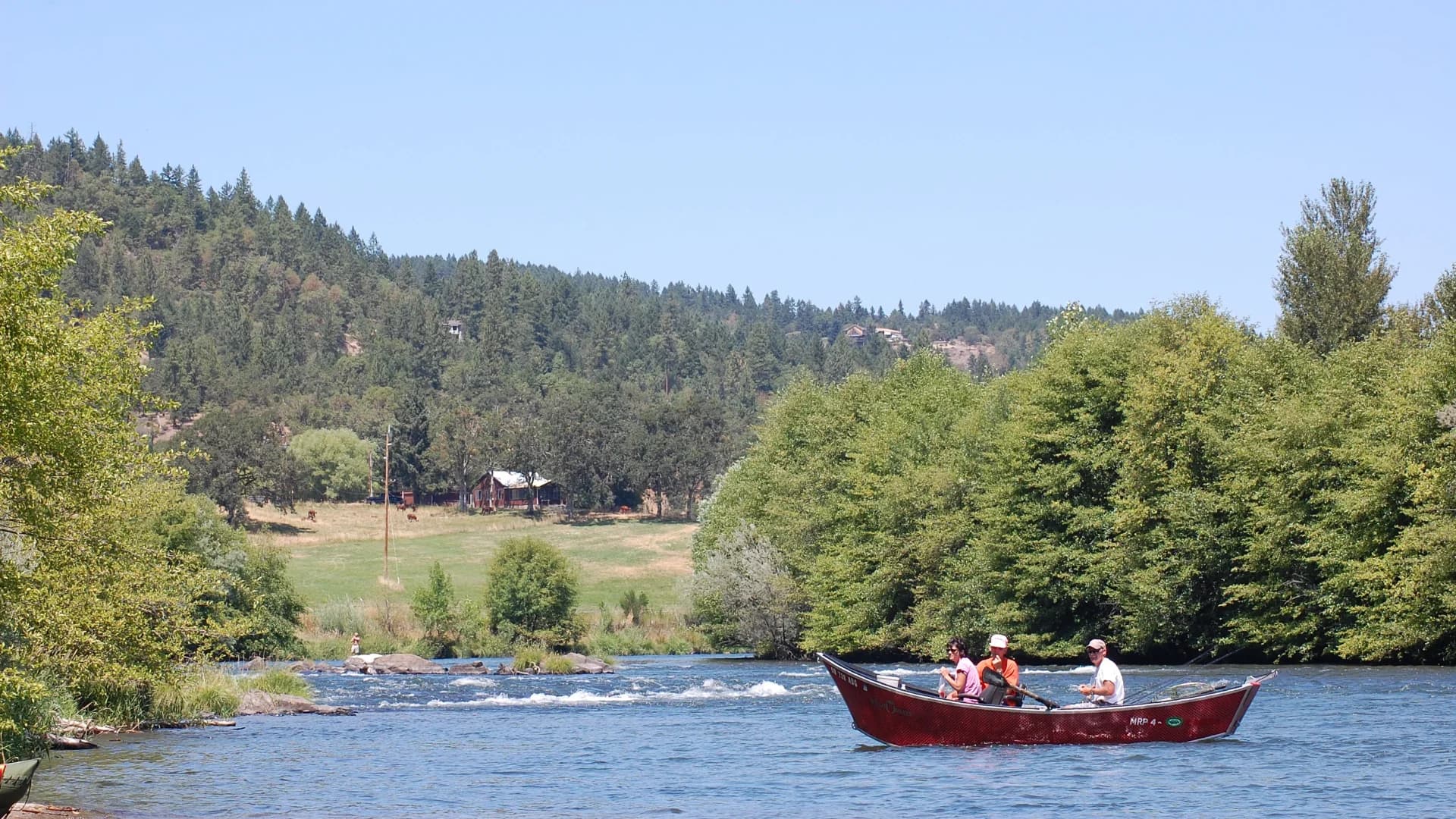 Three people in a red boat navigate a river surrounded by trees and hills on a sunny day.