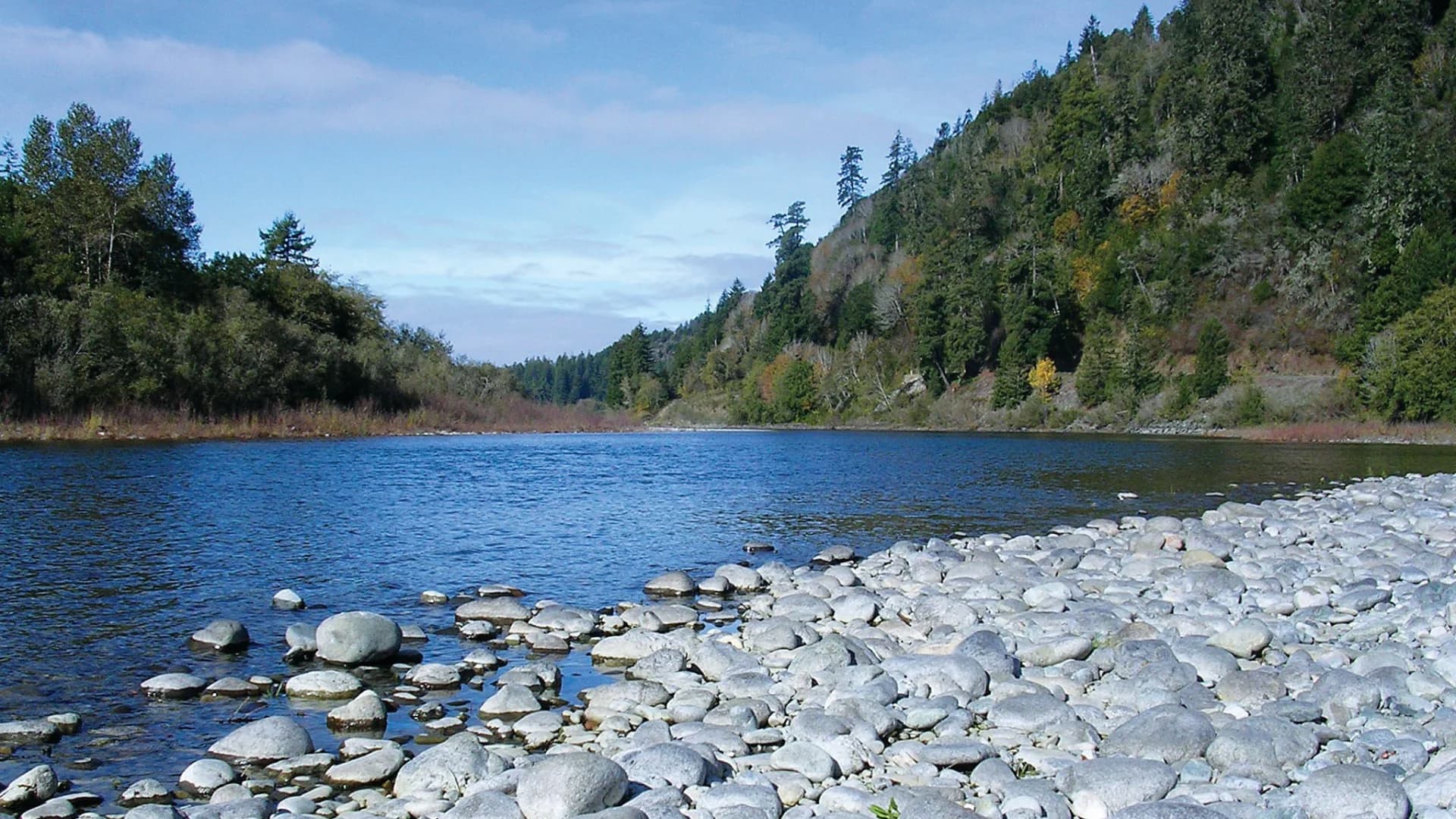 A serene river scene with smooth stones lining the shore, surrounded by lush green hills and a blue sky.