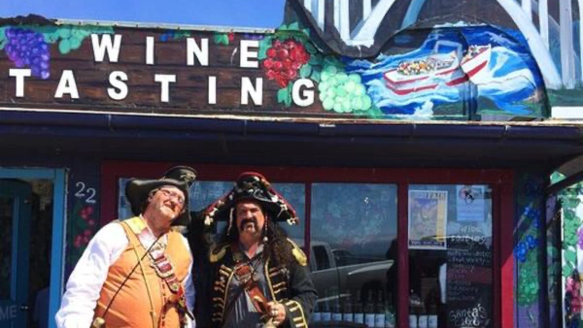 Two men dressed as pirates stand in front of a colorful "Wine Tasting" sign at a wine shop.