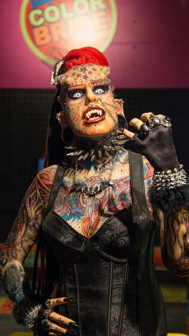 Tattooed individual with red hair and dramatic makeup, wearing a black corset and spiked accessories. Background displays colorful signage.