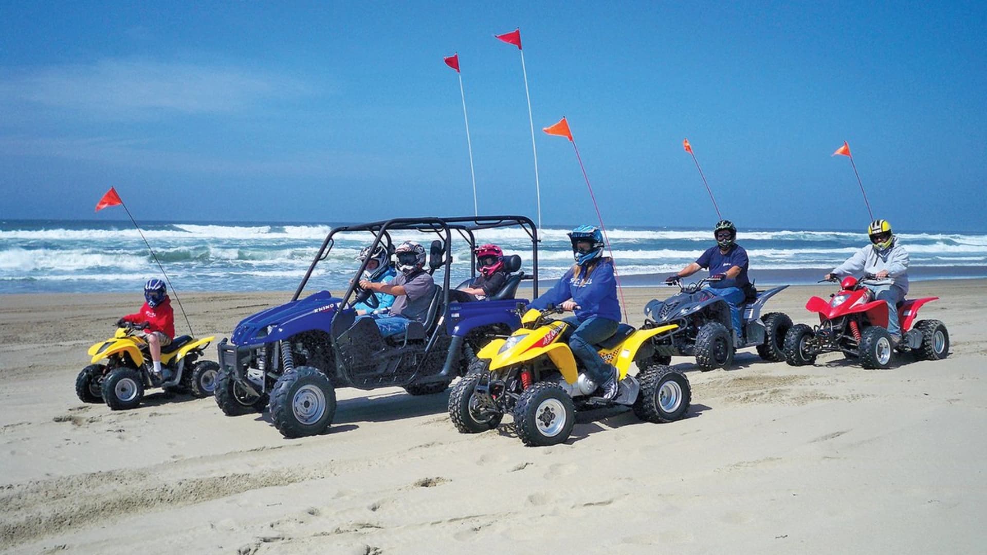 Group of riders on ATVs and a dune buggy on a sandy beach, with ocean waves in the background.