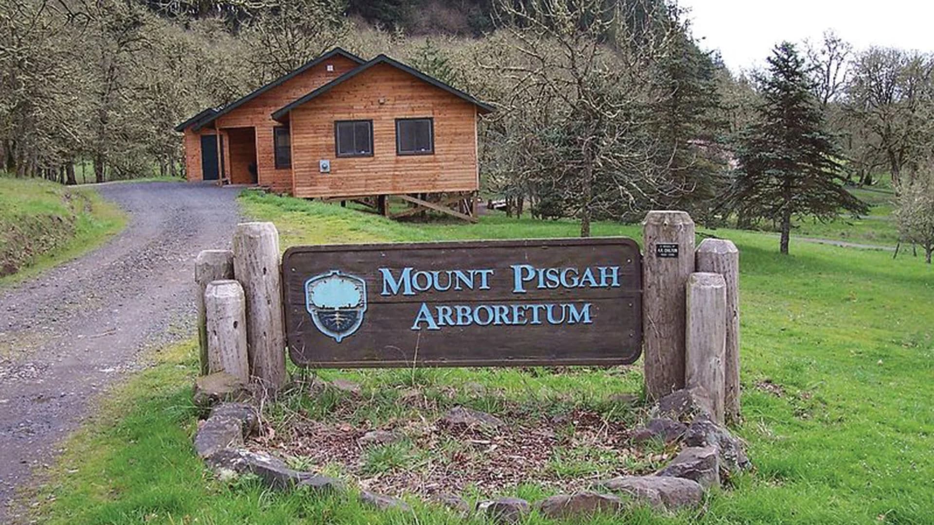 Wooden sign for Mount Pisgah Arboretum near a cabin on a grassy path surrounded by trees.