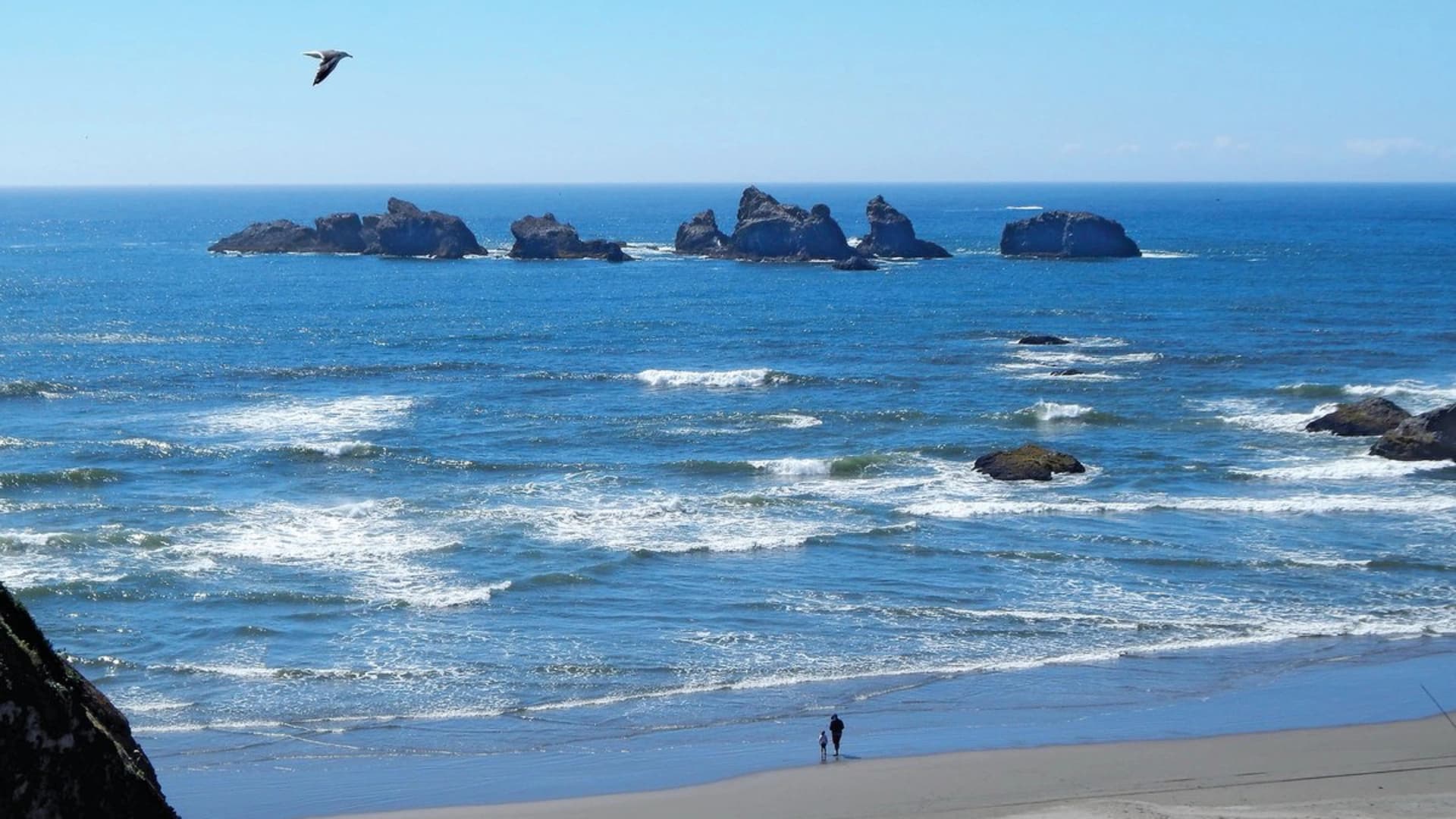 A child and an adult walk on a beach with ocean waves and rocky islets in the distance under a clear blue sky.