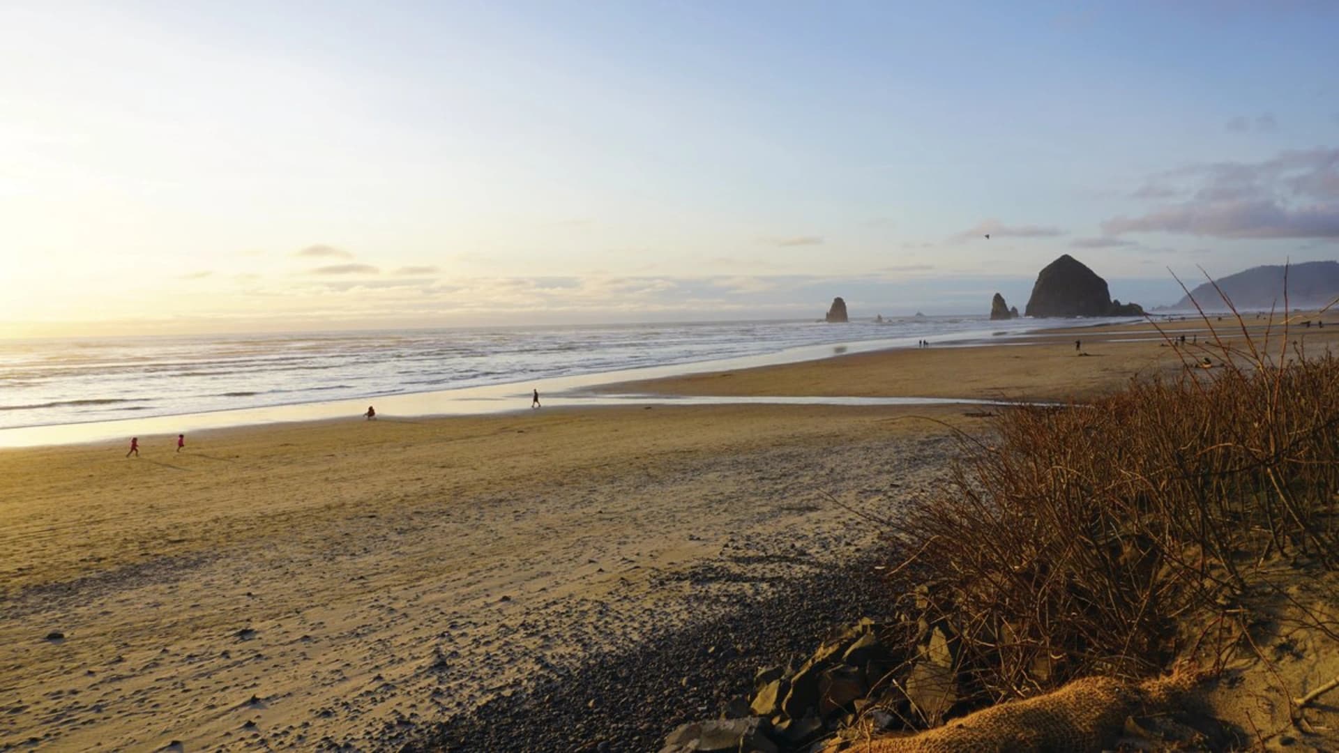 Sunset at a beach with people walking along the shore, rocky formations in the distance.