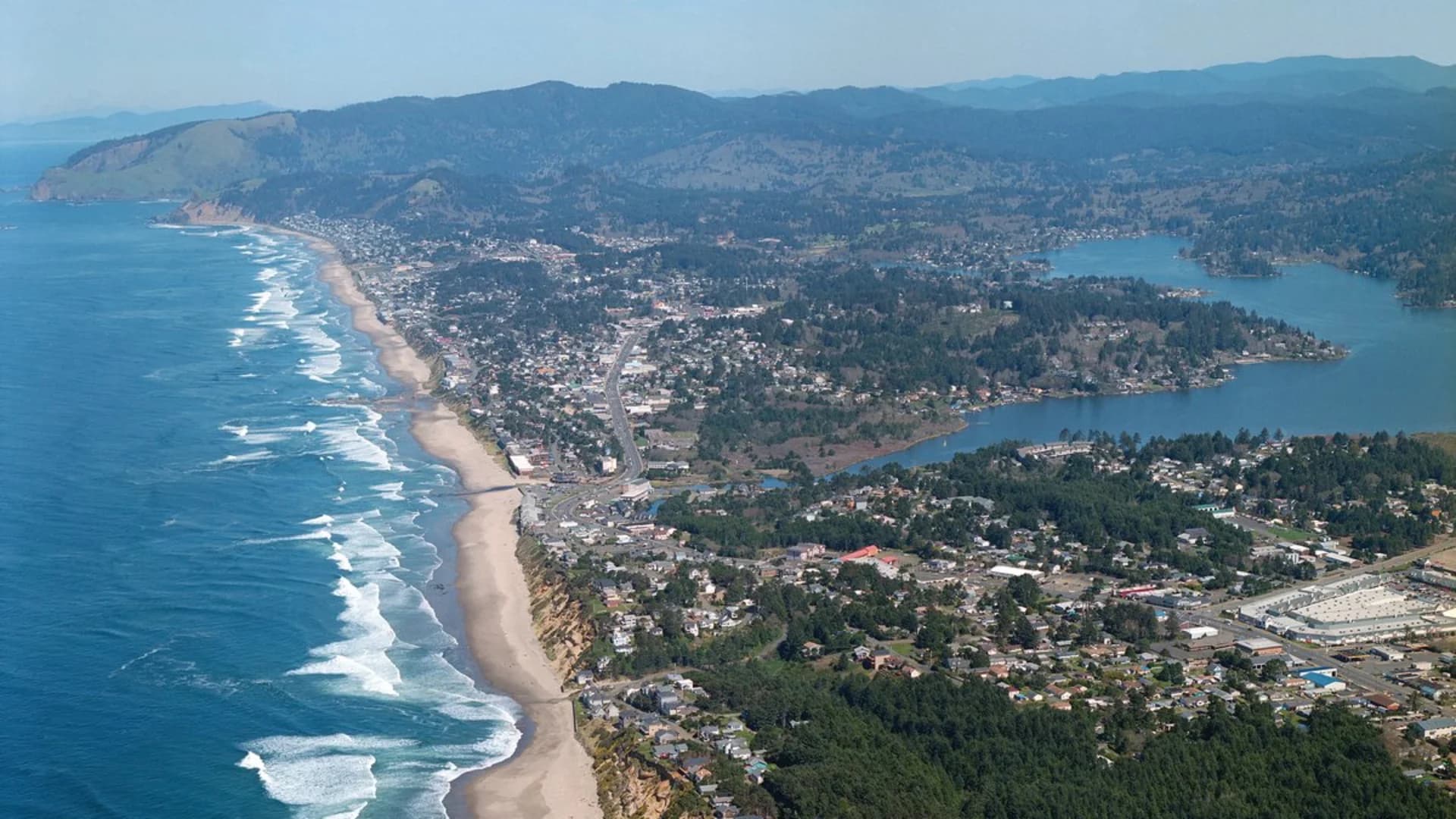 Aerial view of a coastal town with beaches, roads, and hills in the background, near a body of water.