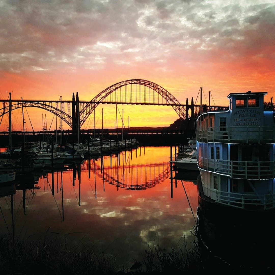 Sunset casts vibrant oranges and purples over a harbor, reflecting the Yaquina Bay Bridge and a docked boat.