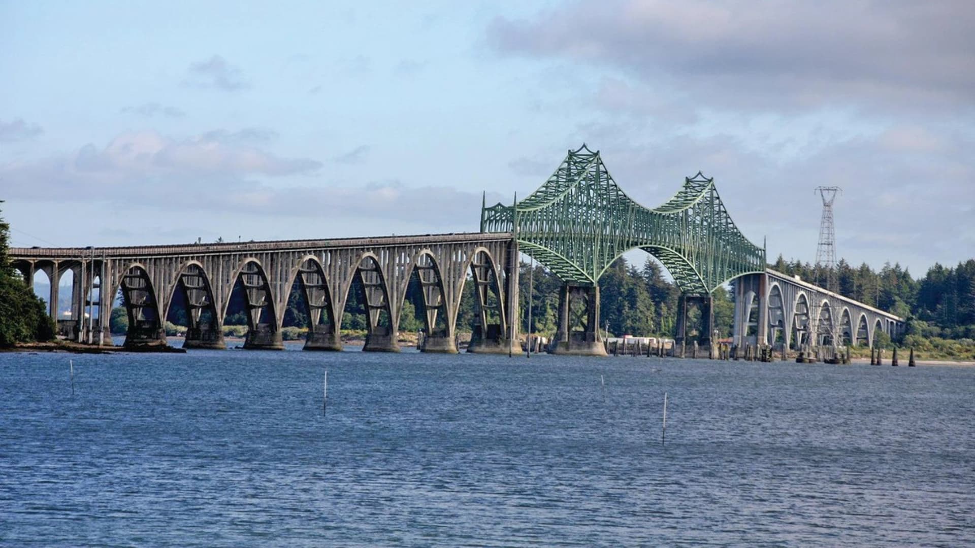A green arch bridge spanning over a body of water, with concrete pillars and a cloudy sky in the background.