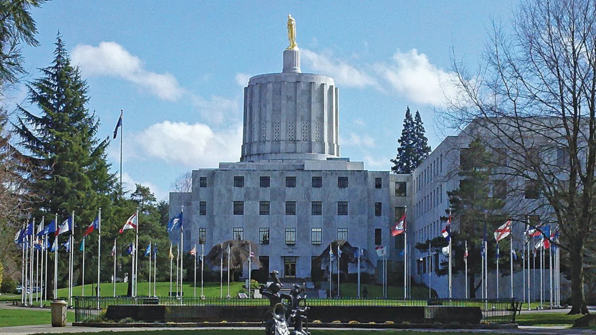 Oregon State Capitol building, featuring a golden statue atop, surrounded by flags and trees against a clear sky.