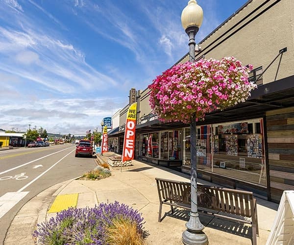 Street view of a storefront with a hanging flower basket and a vertical sign reading “WE’RE OPEN”